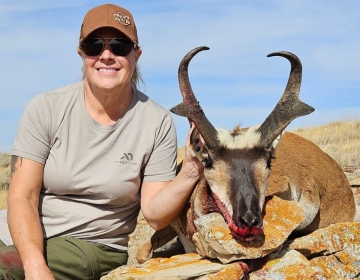 A female hunter wearing a First Lite shirt and sunglasses sits beside a harvested pronghorn antelope resting on a rocky outcrop. The scenic Wyoming terrain enhances the photograph.